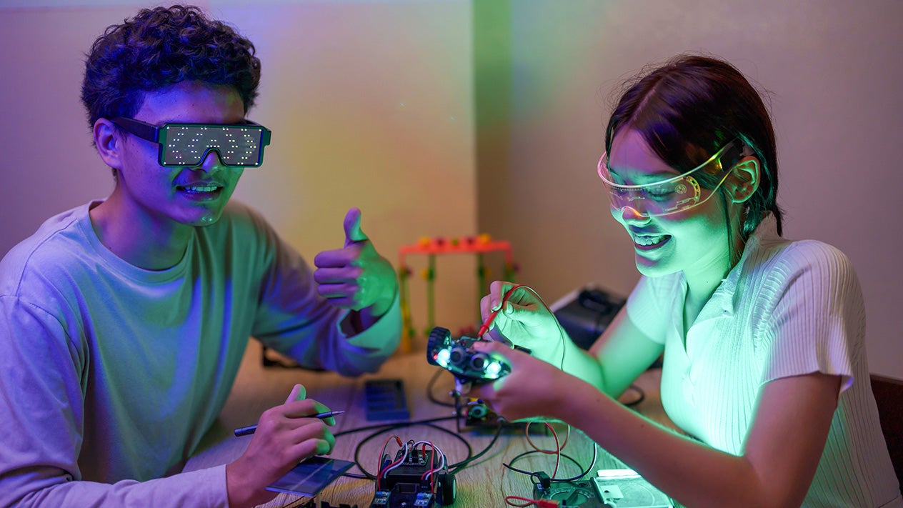 Young man and woman wearing safety goggles, smiling and focused while working on an electronics project with glowing components on a table.