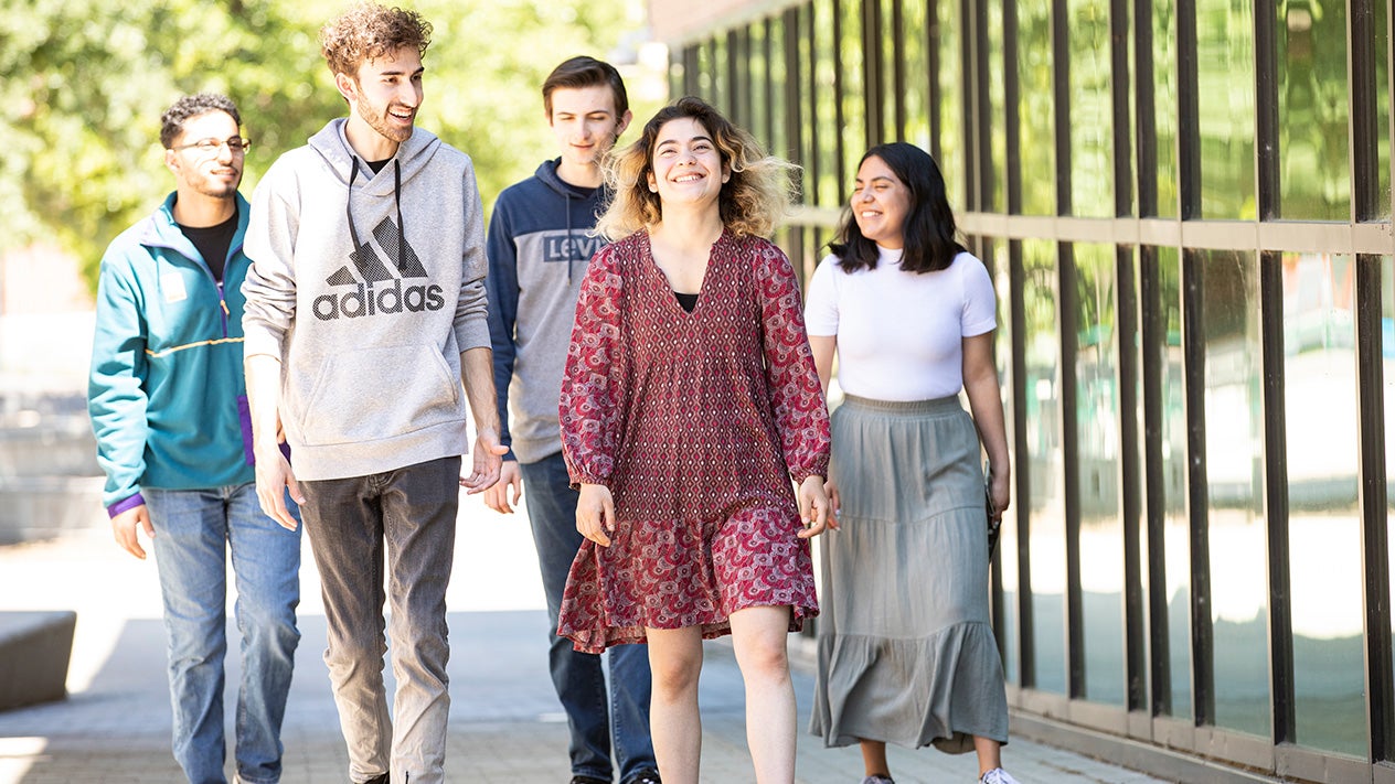 Five students smile and laugh while walking together down a sidewalk.