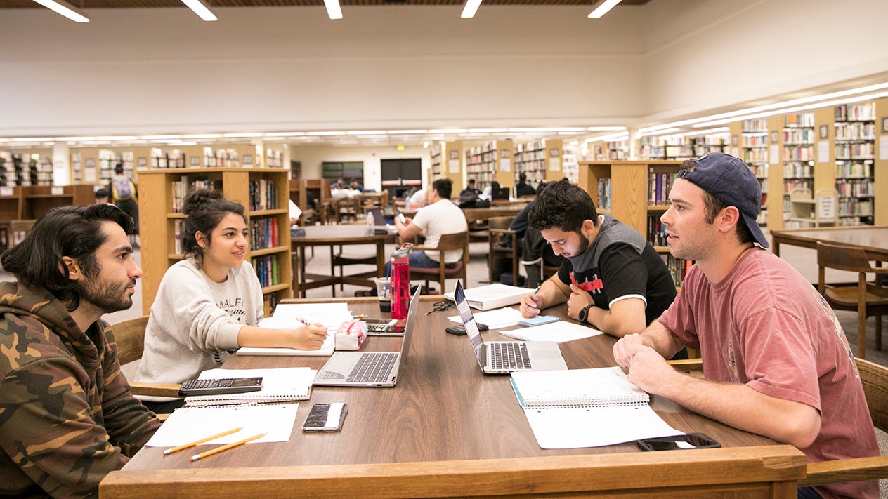 Four students are sitting around a table in a library, studying with laptops and notebooks. The library is spacious with bookshelves and other students in the background.