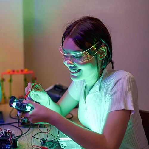 Young woman wearing safety goggles, smiling and focused while working on an electronics project with glowing components on a table.