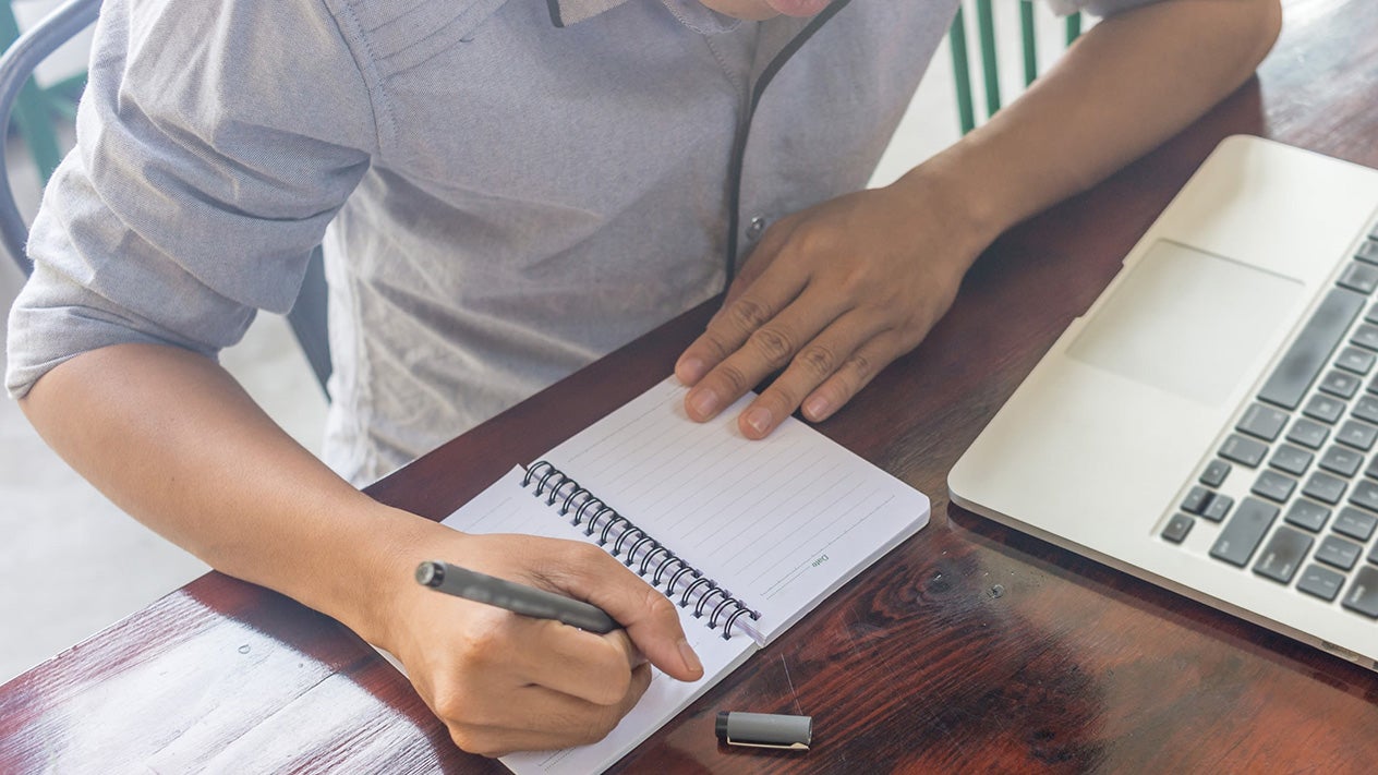 Student writing in notebook next to computer