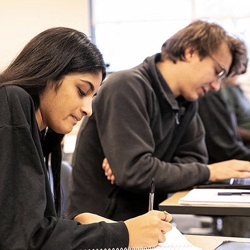 Students in a classroom, writing and typing on computer