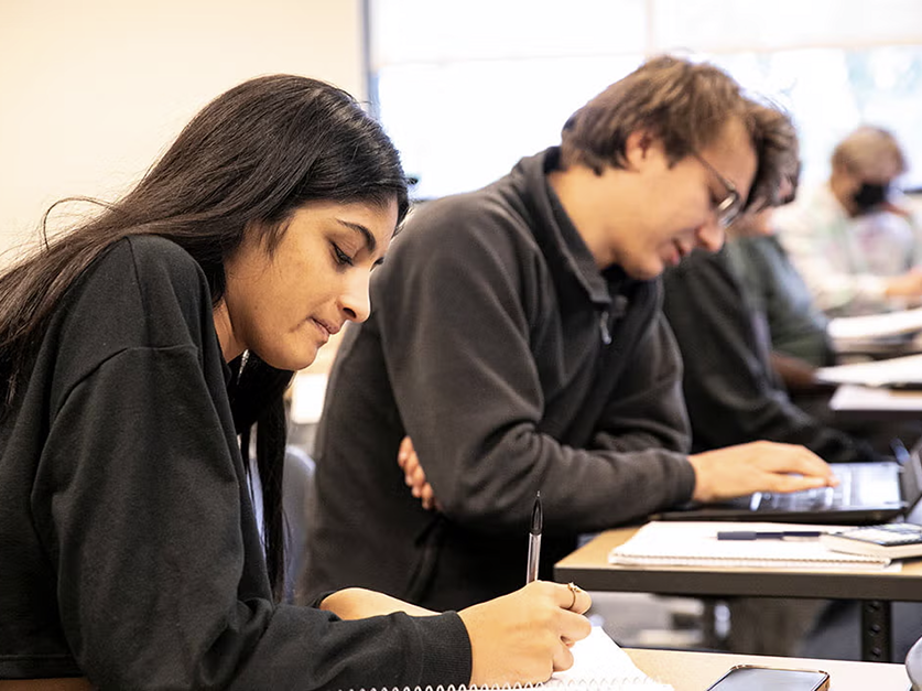 Students in a classroom, writing and typing on computer